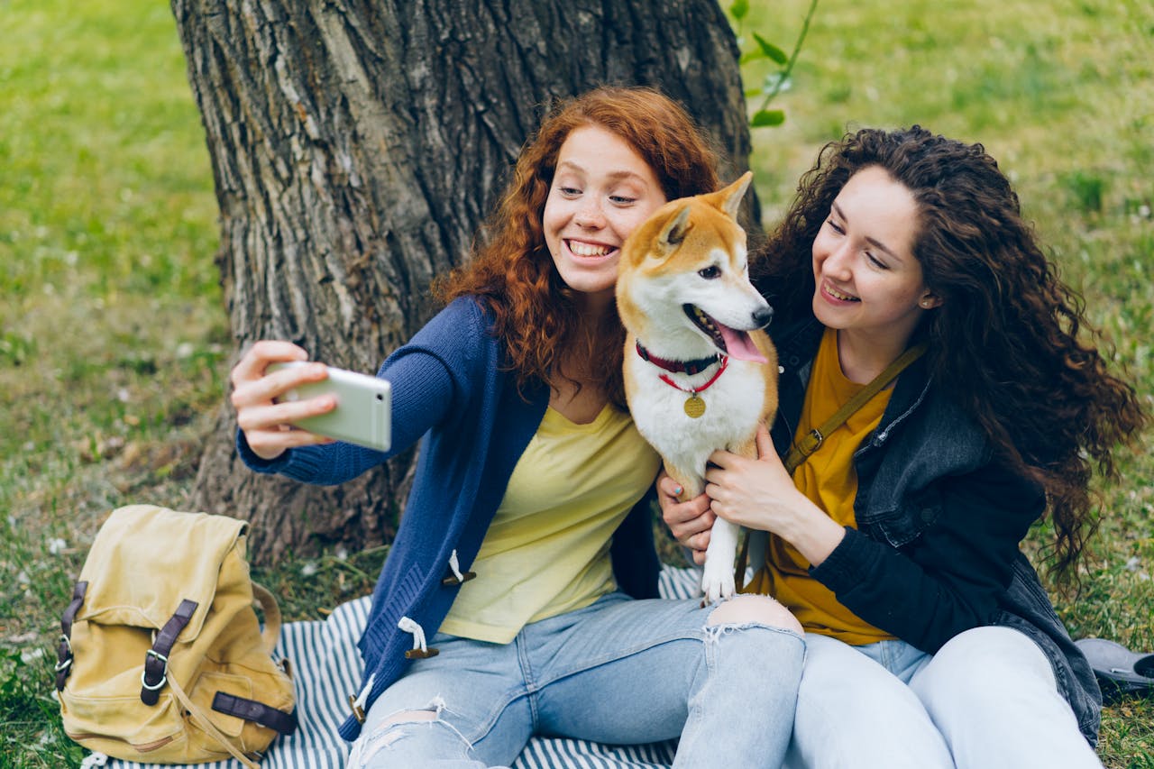 hero-img-02 Two young women smiling while taking a selfie with a Shiba Inu dog in a park setting.